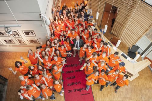 Eine große Gruppe junger Menschen blickt von unten hoch zur Kamera und streckt die Arme hoch.