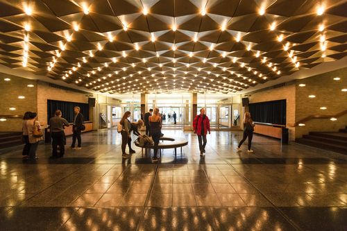 Ein hell erleuchtetes Foyer mit dramatischem Licht, einige Personen bewegen sich durch das Foyer.
