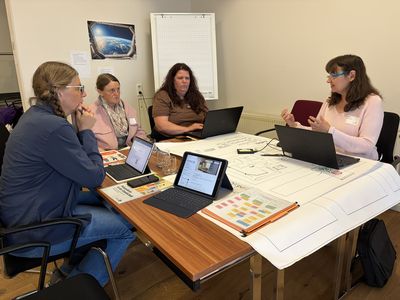 Vier Lehrkräfte sitzen um einen Tisch herum und sprechen miteinander, sie haben Laptops und Tablets vor sich.