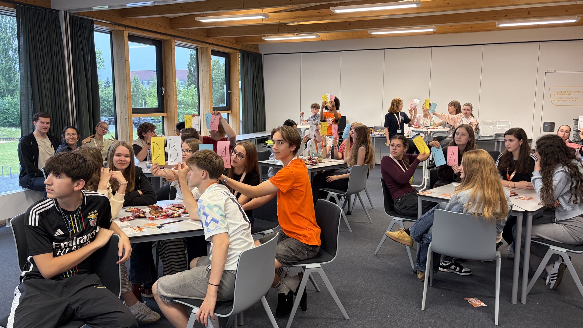 Eine Gruppe Schüler*innen sitzt in einem Konferenzraum, viele halten Antwort-Karten mit Buchstaben darauf in die Höhe.