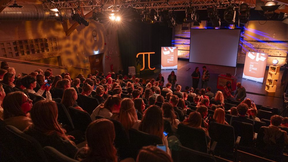 Blick von der obersten Reihe auf ein voll besetztes Theater, auf der Bühne sind Roll-ups, ein Regal mit Globus, eine Leinwand