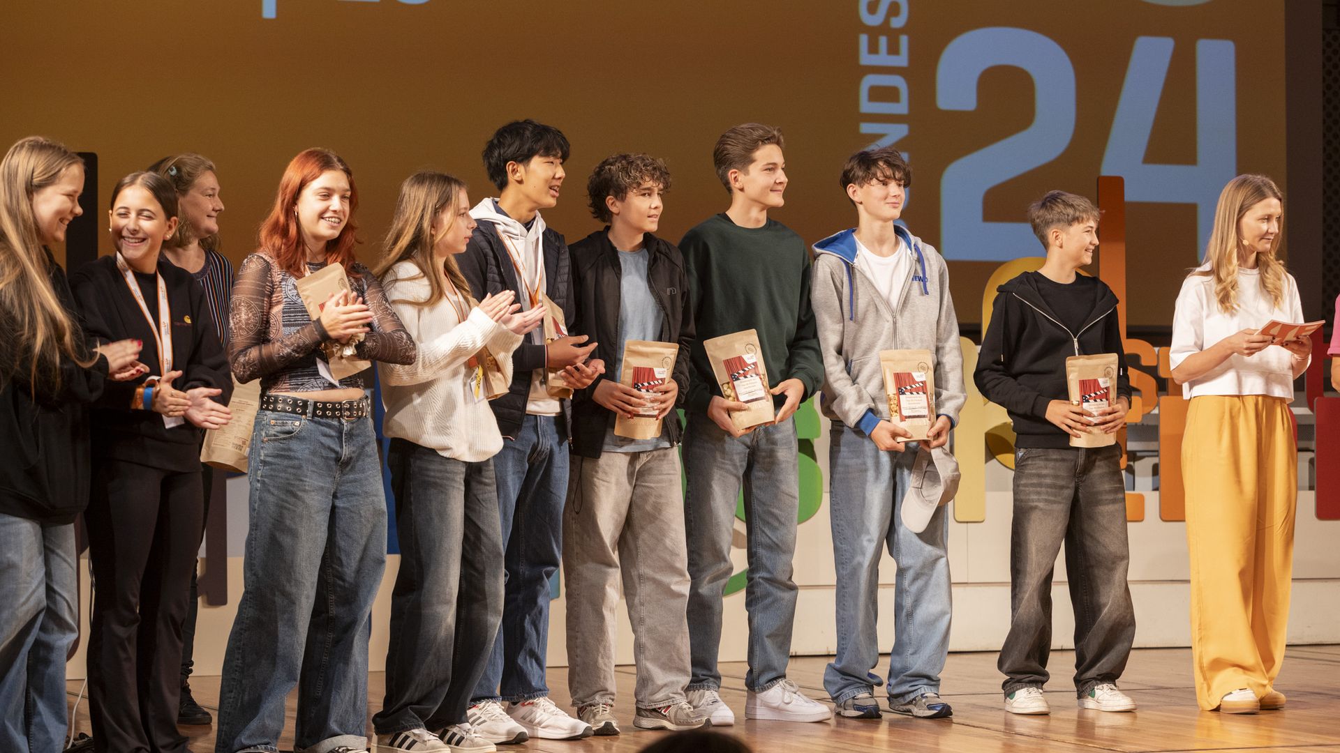 Schülerinnen und Schüler stehen auf der Bühne in einer Reihe und erhalten von der Moderatorin Popcorntüten als Auszeichnung.