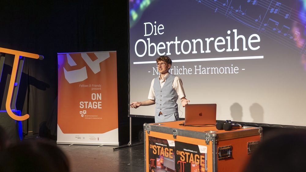 Ein junger Mann präsentiert vor einer großen Leinwand. Im Hintergrund ist ein orangenes Banner zu sehen.