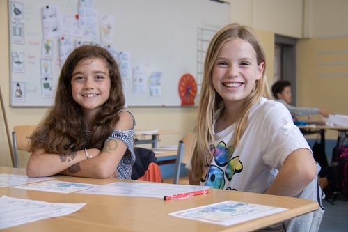 Zwei Grundschülerinnen sitzen an ihrem Schreibtisch im Klassenzimmer und lächeln in die Kamera.