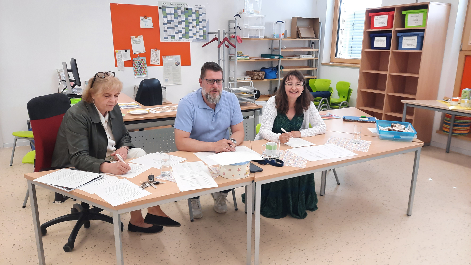 Drei Lehrkräfte sitzen an einem Tisch und lächeln in die Kamera.