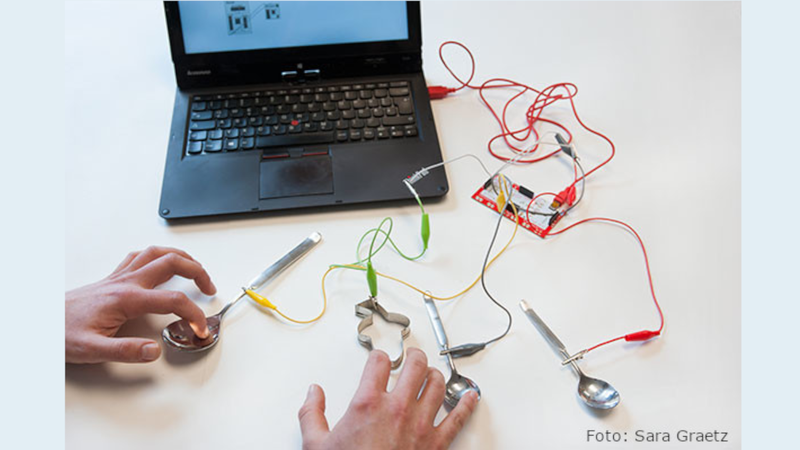 Ein Laptop ist über Kabel und Klammern mit Löffeln verbunden. Zwei Hände positionieren die Gegenstände.