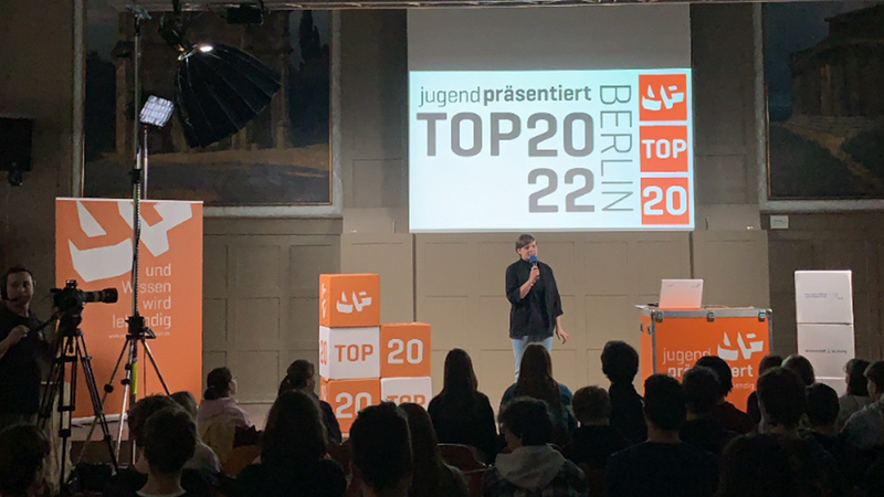 Top-20-Event: Friederike Gräßer moderiert auf einer Bühne vor Publikum. An der Seite steht ein Kameramann und filmt.